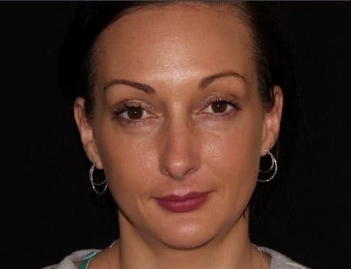 A close-up frontal portrait of a woman with a neutral expression, light skin, and dark hair slicked back. she wears silver hoop earrings and a light grey top.