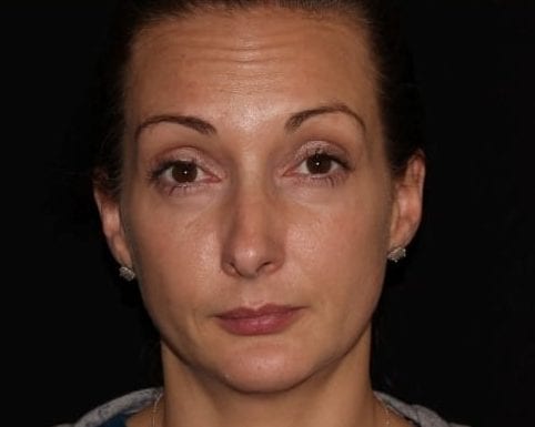 Close-up of a woman with a neutral expression, wearing small earrings, against a black background. she has her hair pulled back and displays minimal makeup.