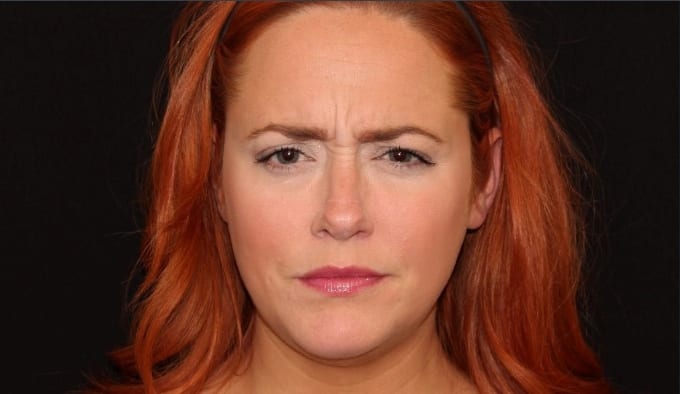 A close-up portrait of a woman with red hair, fair skin, and slight frown, looking at the camera against a black background.