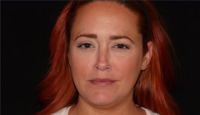 Headshot of a woman with red hair pulled back, clear complexion, and a slight smile, set against a black background. she wears a white shirt and is looking directly at the camera.