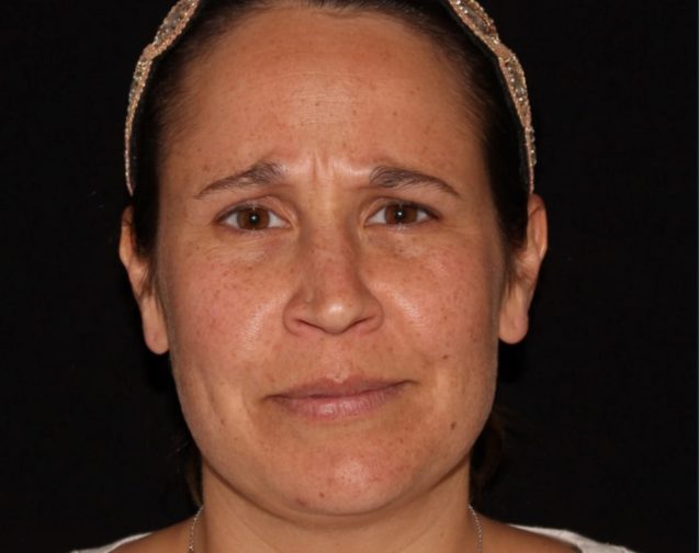 A close-up portrait of a woman with a neutral expression, featuring shoulder-length brown hair held back with a beaded headband, set against a black background.