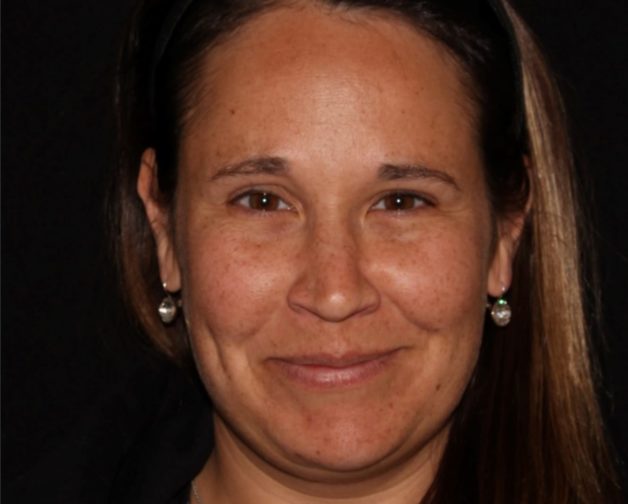 Portrait of a smiling woman with long brown hair pulled back, wearing a black headband and pearl earrings, against a dark background.