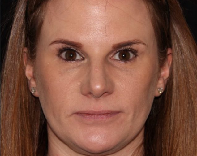 Close-up of a woman with a neutral expression, featuring straight light brown hair pulled back, clear skin, and piercing brown eyes. she wears simple stud earrings.