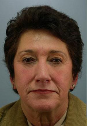 Portrait of an older woman with short brown hair, wearing a beige coat, looking directly at the camera with a neutral expression.