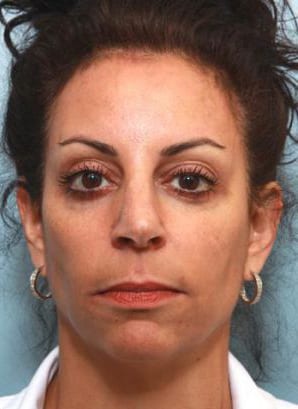 Close-up portrait of a woman with dark hair pulled back, wearing hoop earrings and a white top, looking directly at the camera with a neutral expression.