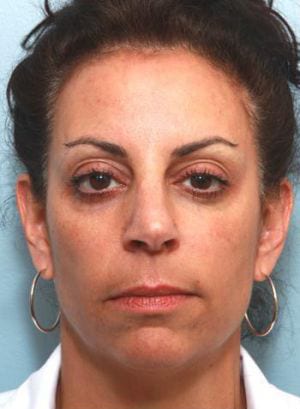 Close-up portrait of a woman with hoop earrings, dark hair pulled back, and minimal makeup, looking straight ahead against a light blue background.