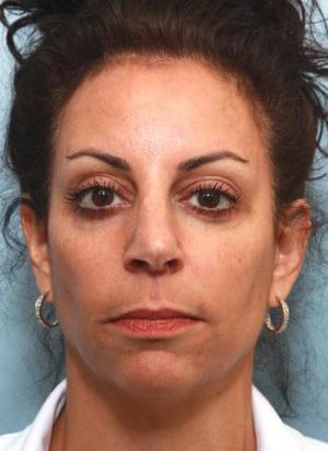 Close-up portrait of a woman with tanned skin, dark curly hair pulled back, wearing makeup and hoop earrings, and a neutral expression against a light blue background.