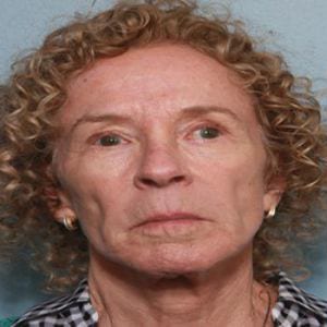 Portrait of an elderly woman with curly, light brown hair, wearing a checkered shirt and small earrings, looking directly at the camera.