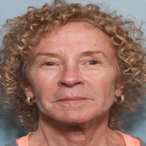 Close-up of a middle-aged woman with curly blonde hair, wearing hoop earrings and an orange top, smiling subtly at the camera.