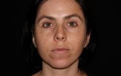 A close-up portrait of a woman with dark hair pulled back, looking directly at the camera with a neutral expression, against a black background.