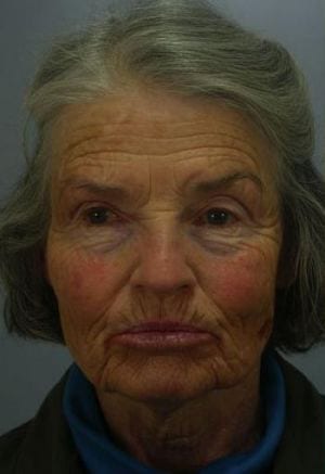 Portrait of an elderly woman with pronounced facial lines, looking directly at the camera, wearing a blue turtleneck. her expression is neutral, and her hair is gray and slightly tousled.