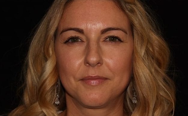 Portrait of a smiling woman with blonde wavy hair and earrings, against a black background.