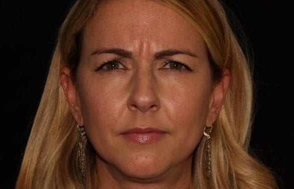 Close-up portrait of a woman with long blonde hair, sporting large hoop earrings, and presenting a serious expression against a black background.