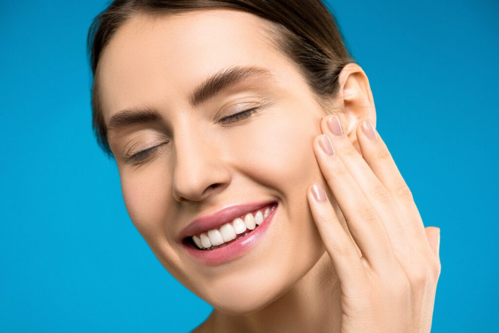 smiling woman wearing light pink lipstick lightly touching her cheekbone