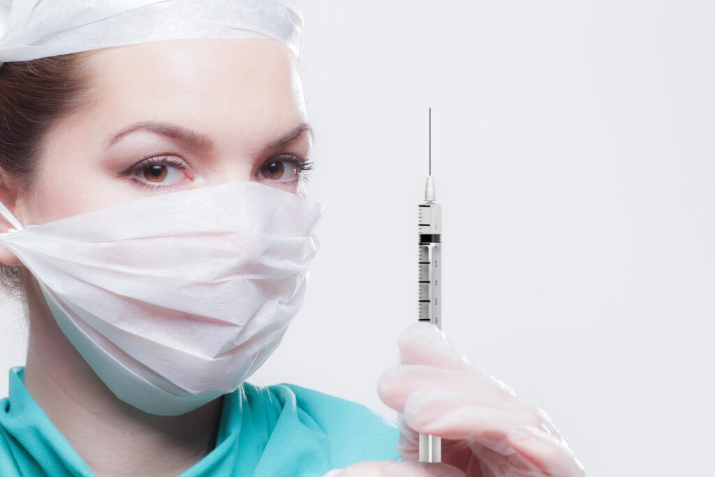 woman wearing medical mask, blue scrubs, and latex gloves holding a needle