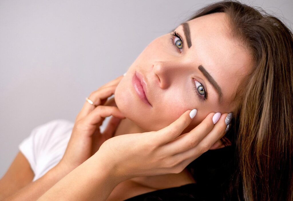 woman with brown hair and light pink nail polish touching her face with her hands