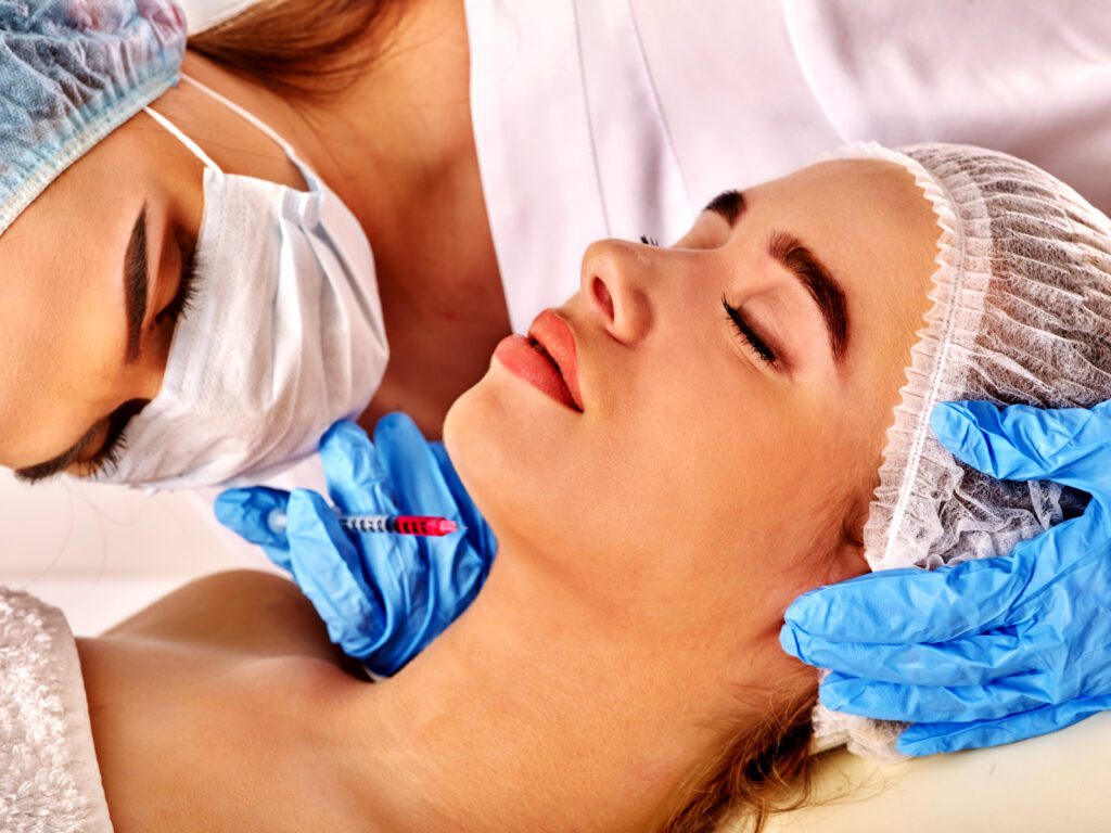 A woman wearing a hair net and a mask while performing a Dysport facial treatment on another woman, who is lying down and relaxing, also in a hair net. Both appear focused and serene.