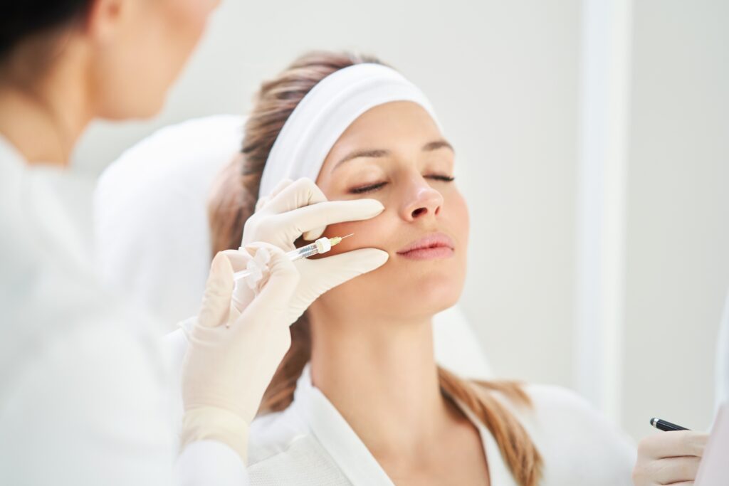 A woman with a headband receiving a facial treatment from a beautician using a tool on her face inquires, "Am I too young for botox?" in a well-lit room.